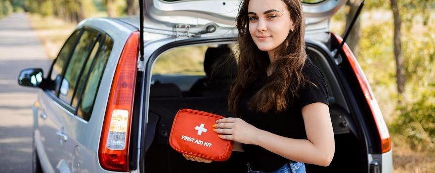 Frau steht am Straßenrand beim Kofferraum ihres Autos und hat den Verbandskasten fürs Auto in der Hand